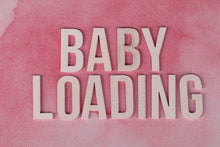 Close-up of 'Baby Loading' text in wooden letters on a pink watercolor background.