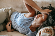 A young woman with curly hair lies indoors, looking distressed and touching her forehead, possibly unwell or suffering from a headache.