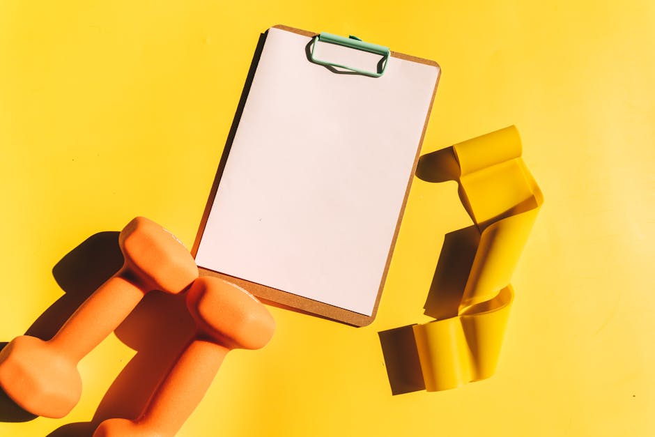Bright flat lay of orange dumbbells, clipboard, and yellow resistance band on yellow background.