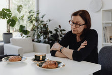 Middle-aged woman in glasses sits at breakfast table with pastries, contemplating.